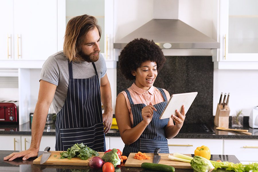 Employee Benefits - A Couple Wearing Matching Striped Aprons are Preparing a Healthy Meal and Checking the Recipe on the Tablet in the Kitchen at Home